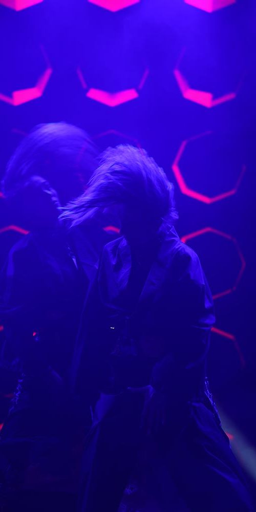 Performer with blurred motion on stage under moody purple lighting and red geometric background visuals.
