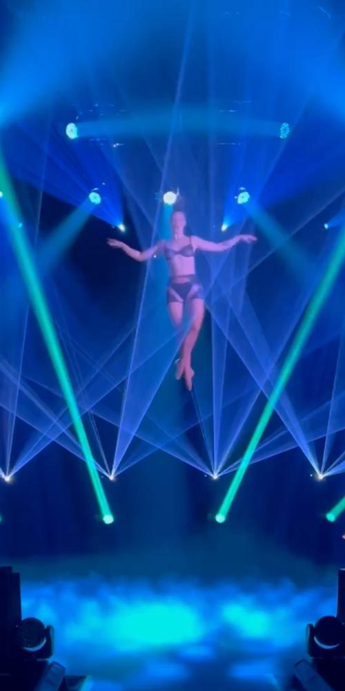 Female performer elevated in mid-air, surrounded by vibrant green and blue lasers in a smoky, high-energy stage show.