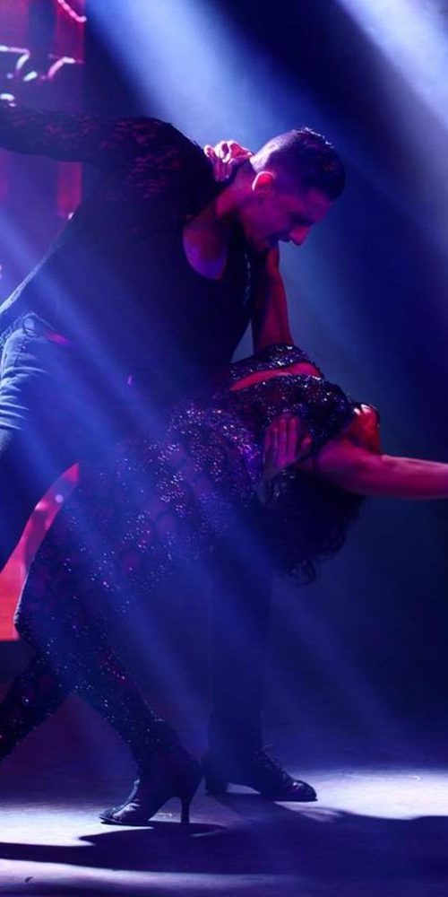 Male and female dance duo in a dramatic ballroom dip on stage, with blue beams of light cutting through a red-themed backdrop.