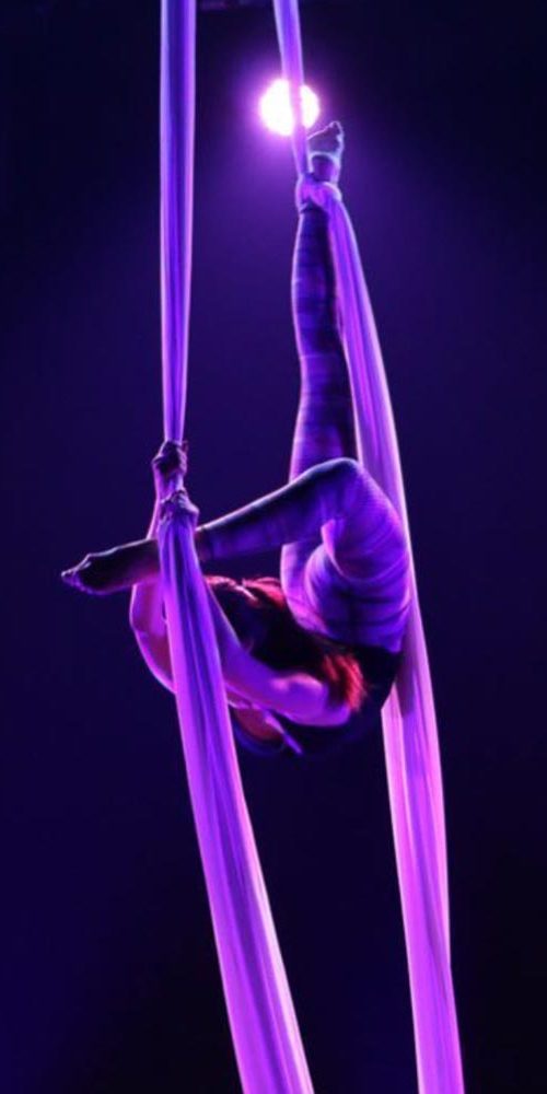 Performer suspended mid-air in an inverted split pose on aerial silks, under focused purple stage lighting.
