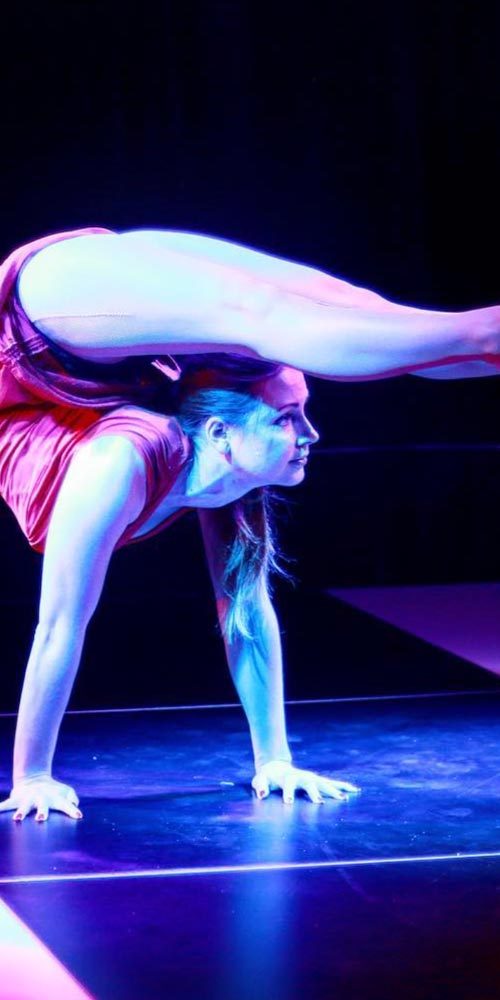 Female contortionist balancing in a handstand with legs arched over head, illuminated by bold pink and blue stage lights.