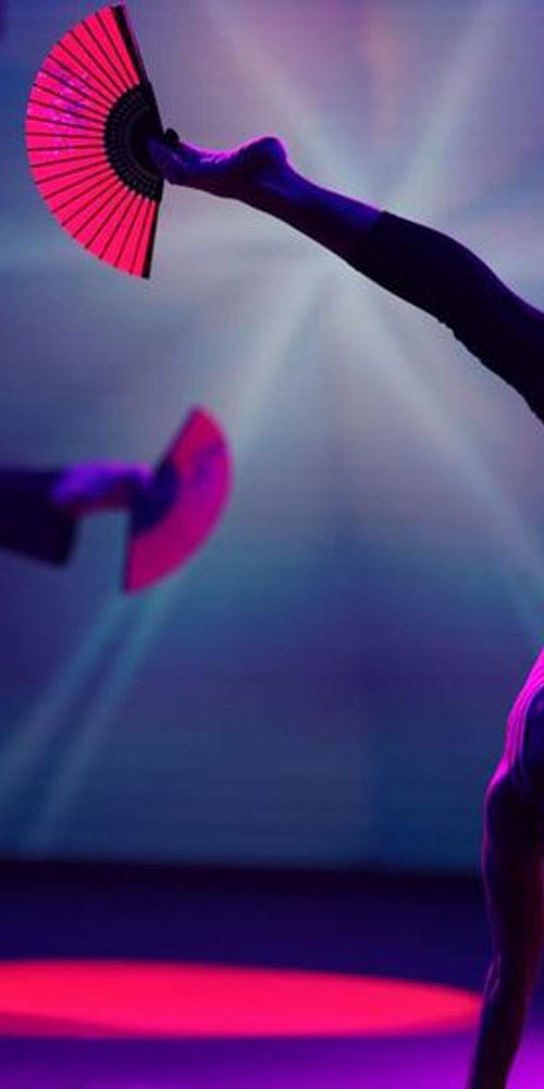 Two performers in simultaneous handstands, each holding a bright pink fan, under dramatic purple-blue stage lighting.