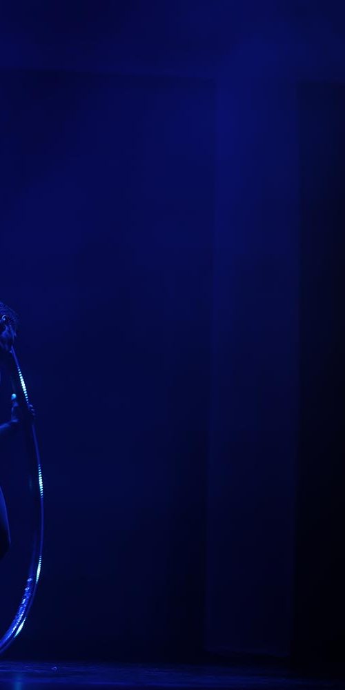 Male acrobat performing inside a large illuminated Cyr wheel on a dark stage with focused blue lighting.