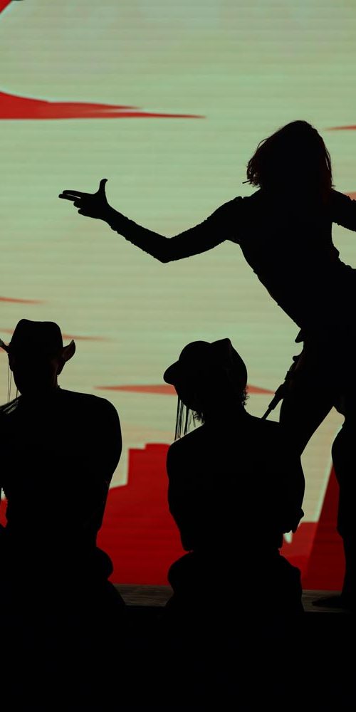 Silhouetted performers posed in front of a red desert-themed LED screen featuring a large setting sun and western landscape.