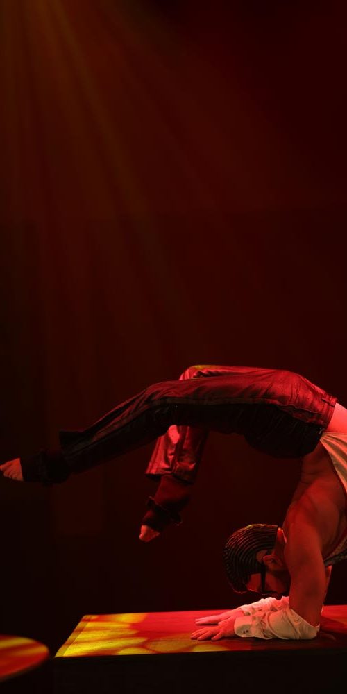 Male contortionist in spotlight balancing in a headstand pose with dramatic backlighting and a red-toned stage atmosphere.