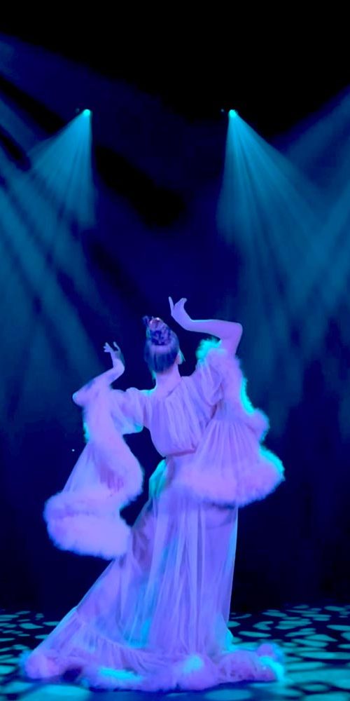 Elegant female performer in flowing white costume dancing solo under sharp blue stage lights with a dark theatrical background.