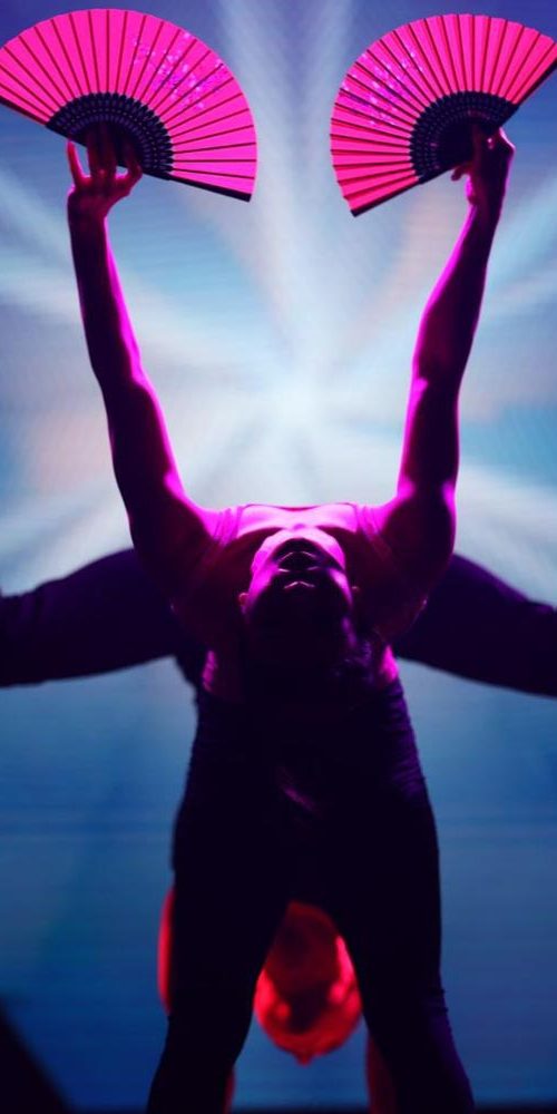 Acrobatic performer holding pink fans while balancing upside down, creating a symmetrical pose under dynamic blue and magenta lighting.