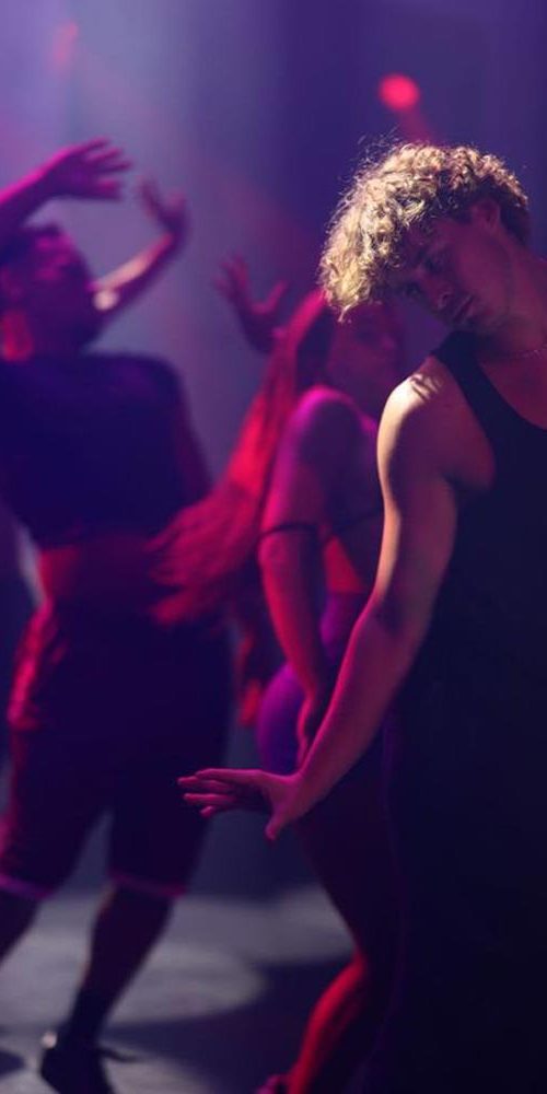 Group of dancers mid-movement in a moody purple-lit performance space, with a male dancer featured in the foreground.