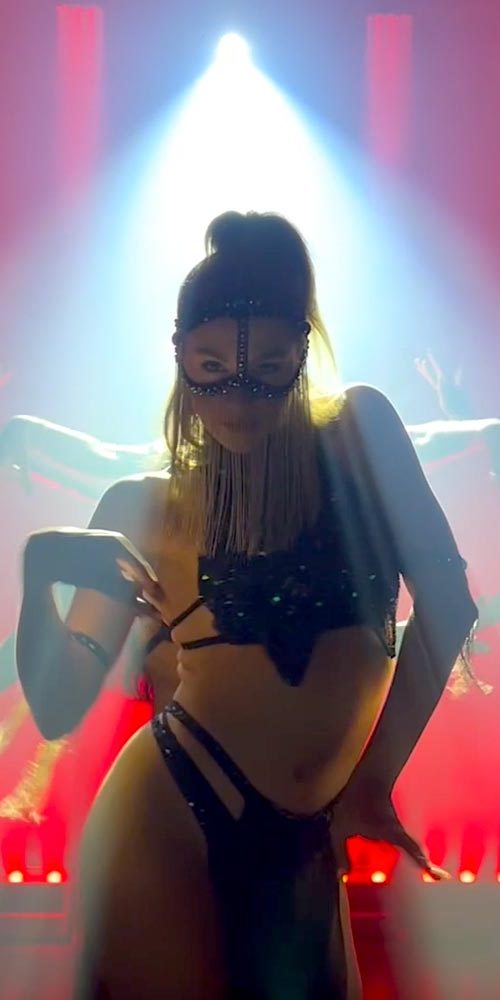 Female performer in black costume and face mask illuminated by spotlight, with backup dancers dramatically posed behind her under red stage lights.