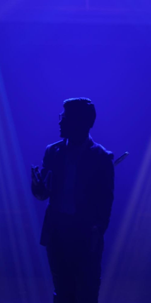Male silhouette standing under dramatic blue stage lighting beams in a minimalist, atmospheric setting.